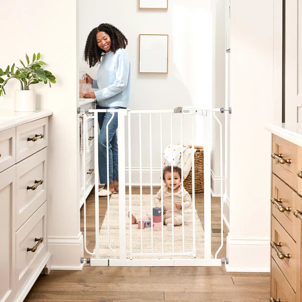 White baby safety gate installed in a home doorway, separating a toddler from the kitchen area to create a secure and baby-proofed living space.
